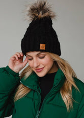 Black diamond cable knit pom hat with natural faux fur pom. Fleece lined for extra warmth and comfort. Classic 100% acrylic design for timeless winter style.