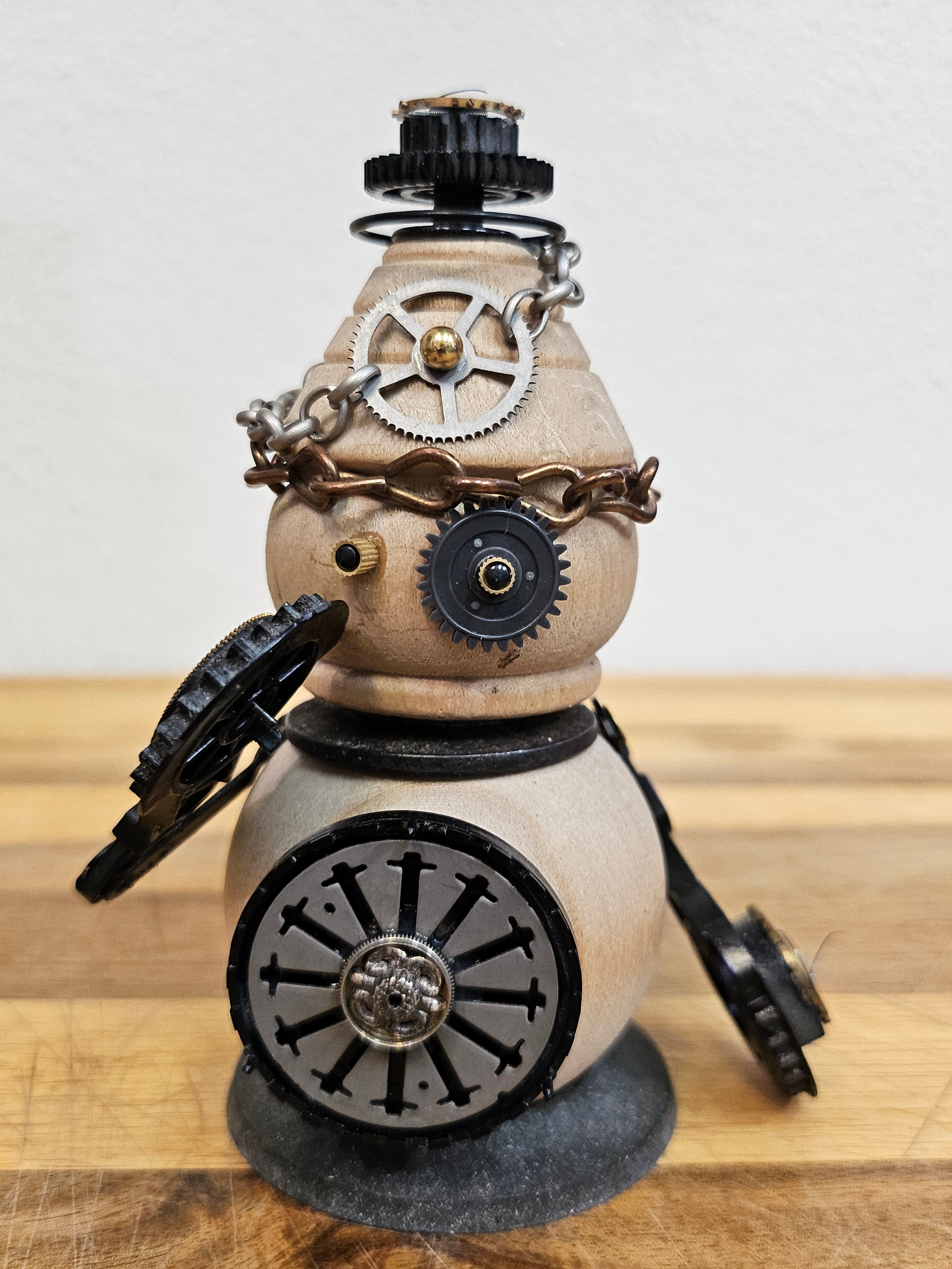 The Handmade Wood Block Steampunk Robot is a unique, decorative figurine crafted from wood with an aged, rustic look. Each robot is one-of-a-kind for display only. Includes a heart-shaped balloon. Available in various sizes, it&