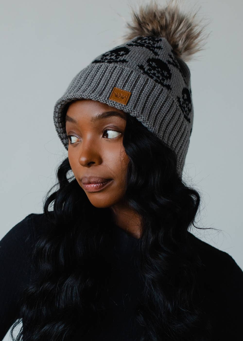 Gray skull pattern knit hat with natural faux fur pom. Fleece lined for extra warmth and style. Crafted from 100% acrylic for a cozy winter essential.