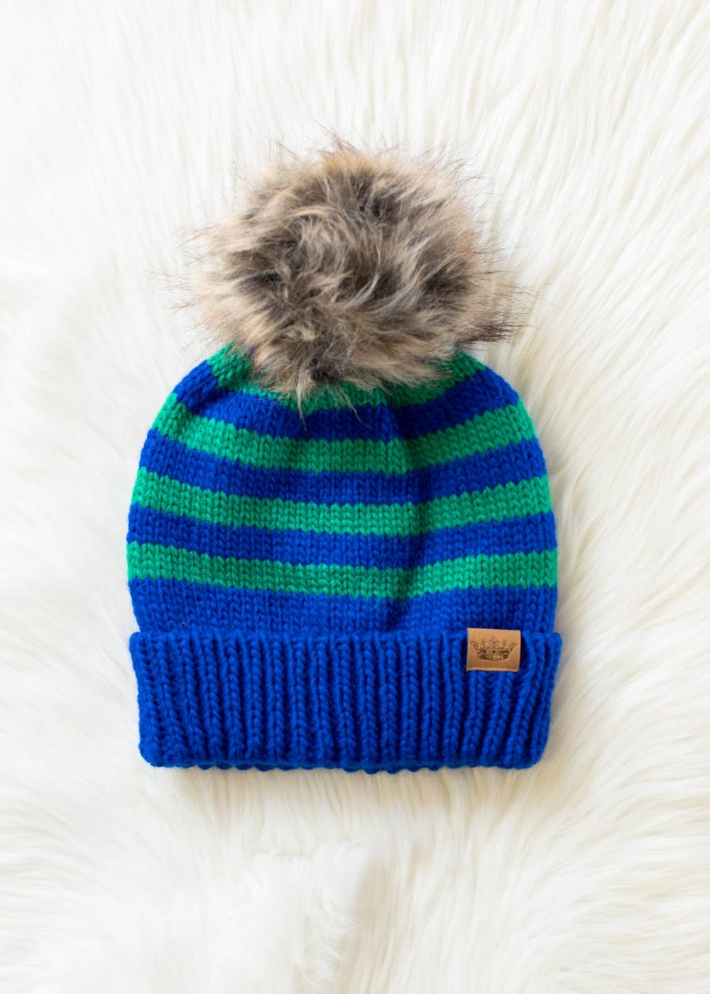 Royal blue and green striped knit hat with a natural-toned faux fur pom and cozy fleece lining. Made from 100% acrylic for warmth and colorful winter style.
