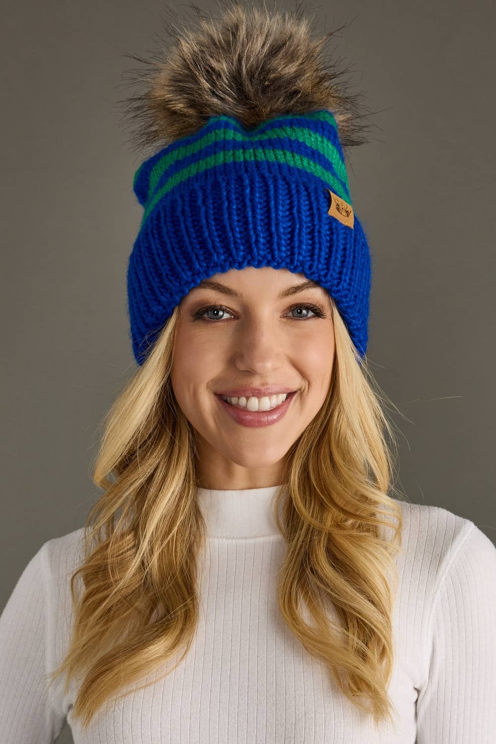 Royal blue and green striped knit hat with a natural-toned faux fur pom and cozy fleece lining. Made from 100% acrylic for warmth and colorful winter style.
