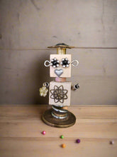 The Handmade Wood Block Steampunk Robot is a unique, decorative figurine crafted from wood with an aged, rustic look. Each robot is one-of-a-kind for display only. Includes a heart-shaped balloon. Available in various sizes, it&