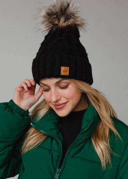Black diamond cable knit pom hat with natural faux fur pom. Fleece lined for extra warmth and comfort. Classic 100% acrylic design for timeless winter style.