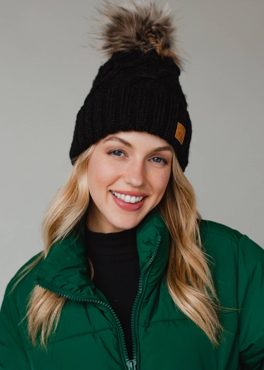 Black diamond cable knit pom hat with natural faux fur pom. Fleece lined for extra warmth and comfort. Classic 100% acrylic design for timeless winter style.