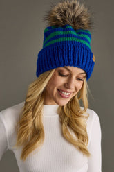 Royal blue and green striped knit hat with a natural-toned faux fur pom and cozy fleece lining. Made from 100% acrylic for warmth and colorful winter style.