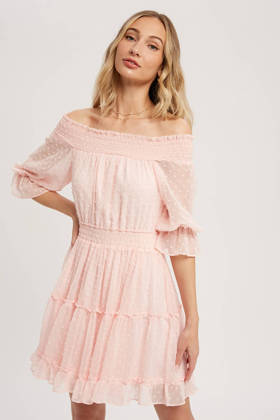 Swiss dot chiffon off-the-shoulder mini dress with elastic waist, bell sleeves, and tiered ruffle hem. Soft blush, relaxed fit, feminine silhouette.