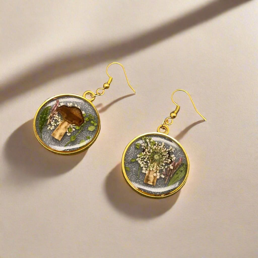 Whimsical dangle earrings with real pressed flowers and tiny mushrooms in resin—each pair is unique and framed in alloy with stainless steel hooks.
