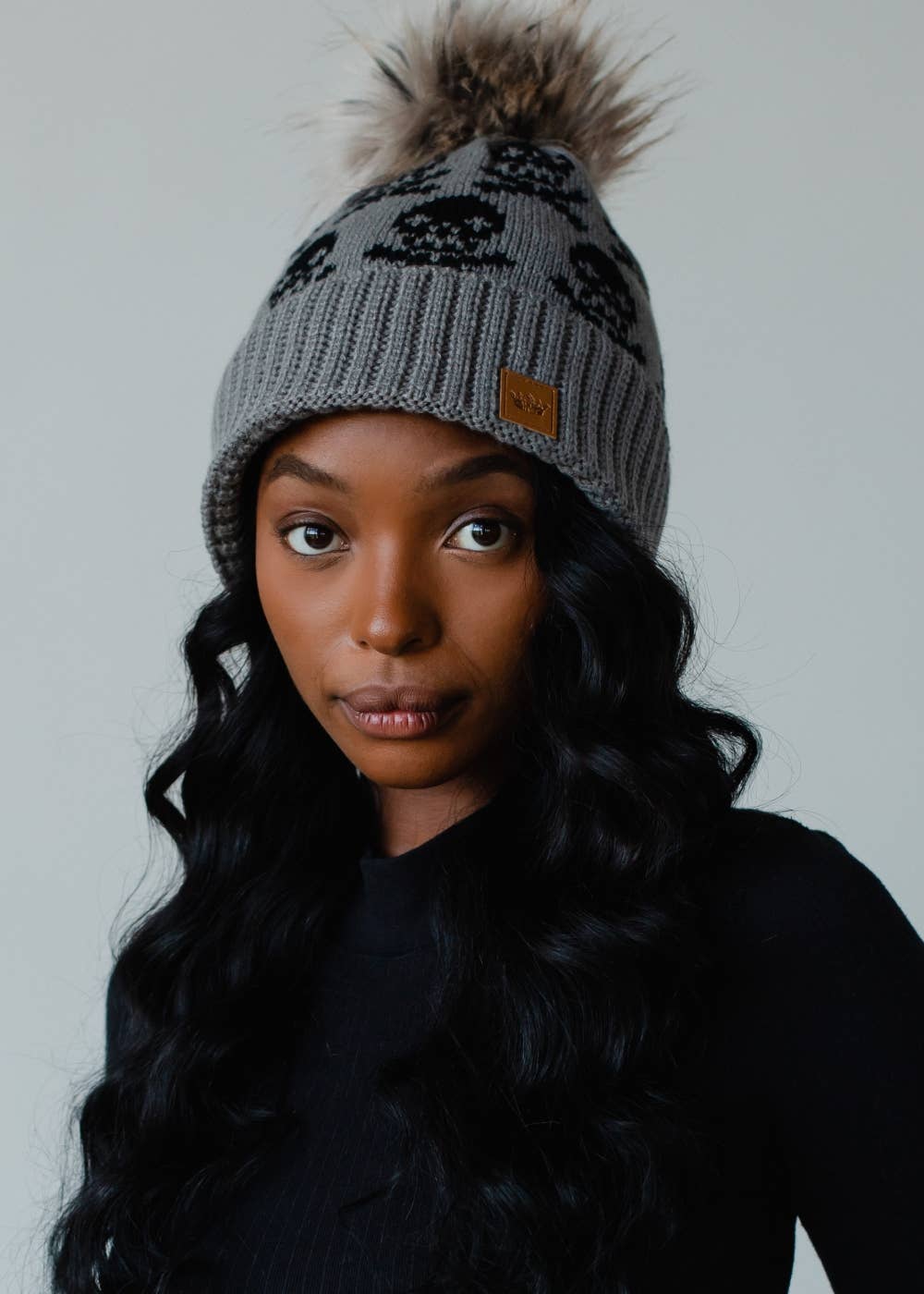 Gray skull pattern knit hat with natural faux fur pom. Fleece lined for extra warmth and style. Crafted from 100% acrylic for a cozy winter essential.