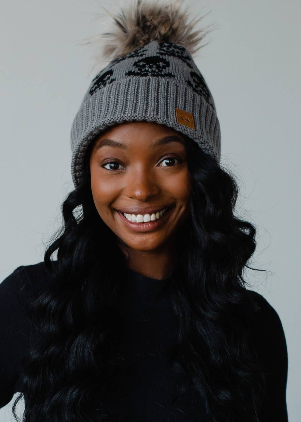Gray skull pattern knit hat with natural faux fur pom. Fleece lined for extra warmth and style. Crafted from 100% acrylic for a cozy winter essential.