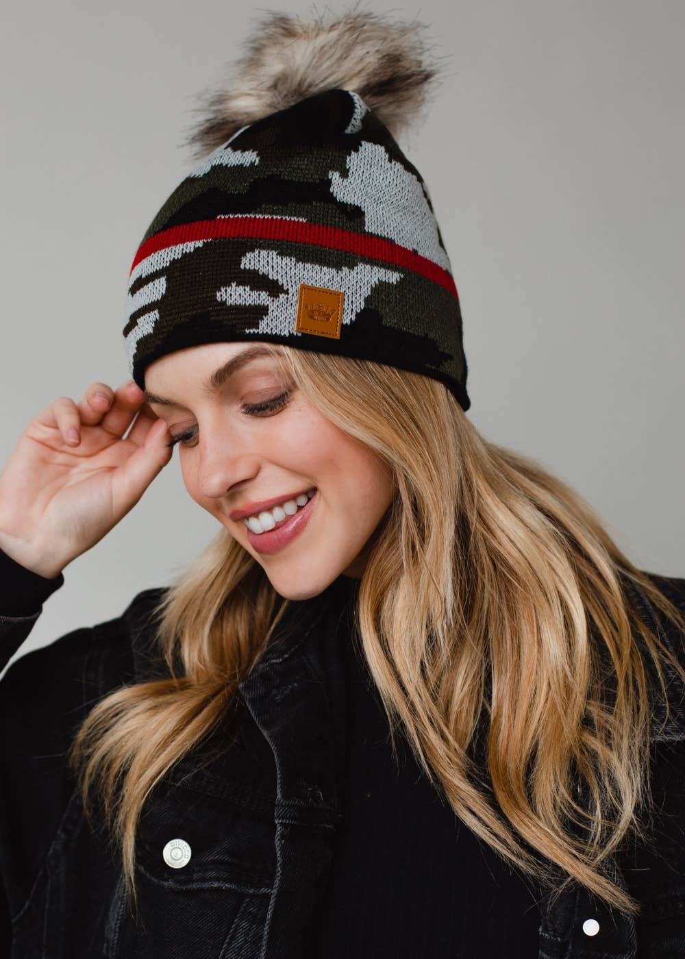 Green camo knit pom hat with bold red stripe detail, a natural-toned faux fur pom and cozy fleece lining. A playful winter accessory for chilly days.