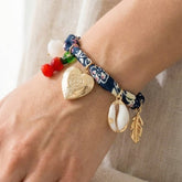 Heart Locket & Charms Bandana Bracelet featuring a heart locket, cowrie shell, feather, and seaside-inspired charms on a vintage-style fabric band.