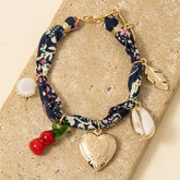 Heart Locket & Charms Bandana Bracelet featuring a heart locket, cowrie shell, feather, and seaside-inspired charms on a vintage-style fabric band.
