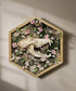 Desert Coyote by Ryan Doran is a 12.5" mixed-media art piece featuring a coyote skull surrounded by preserved pink wildflowers in an octagonal wooden frame. This striking installation blends desert resilience with delicate natural beauty, making it a captivating addition to any space.