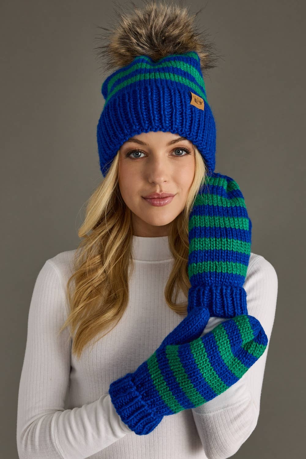 Royal blue and green striped knit hat with a natural-toned faux fur pom and cozy fleece lining. Made from 100% acrylic for warmth and colorful winter style.