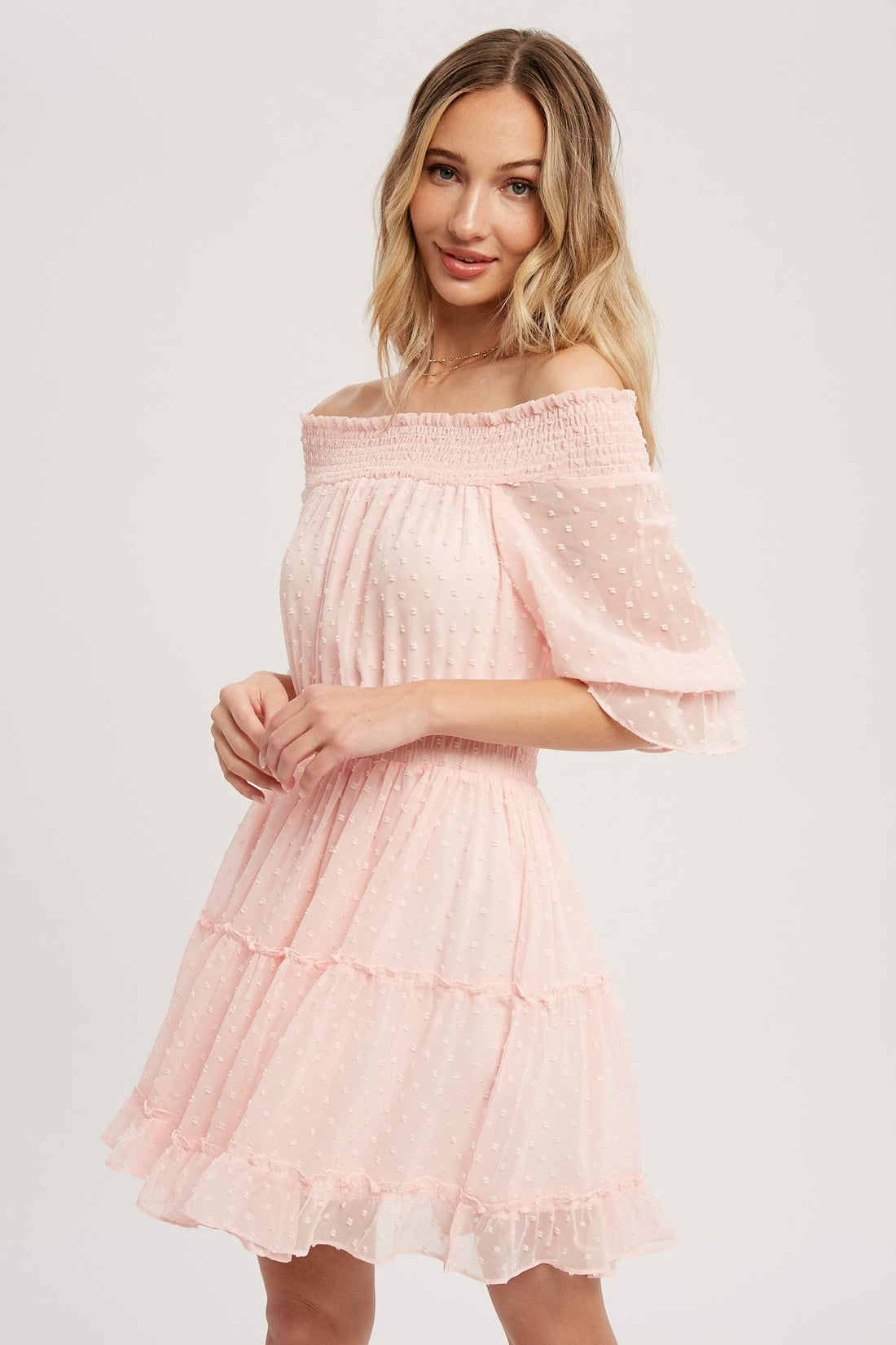 Swiss dot chiffon off-the-shoulder mini dress with elastic waist, bell sleeves, and tiered ruffle hem. Soft blush, relaxed fit, feminine silhouette.